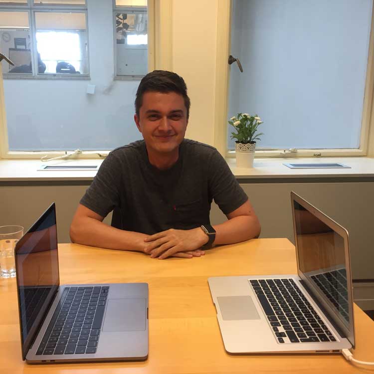 Man (Mlevin from Onesix) sitting at a table with two open laptops facing each other, smiling, in a room with a window and a potted plant behind him.